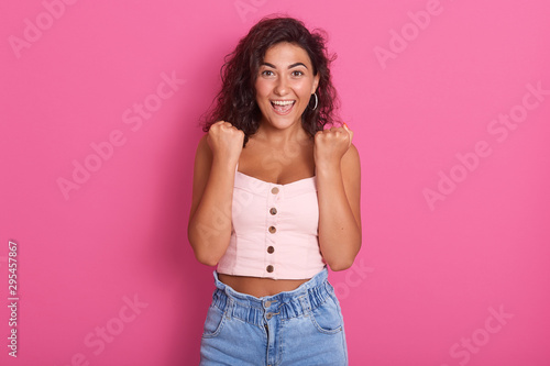 Photo of happy young woman with beautiful dark wawy hair screaming and clenching fist while looking directlyat camera, wearing stylish outfit, isolated over pink background. People emotions concept. © sementsova321