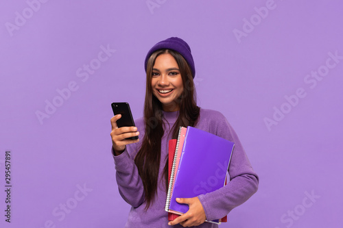 Pretty african-american teenager student in a hat and sweater holding folders and looking surprised checking notification on a smartphone standing isolated over purple background. Concept of education