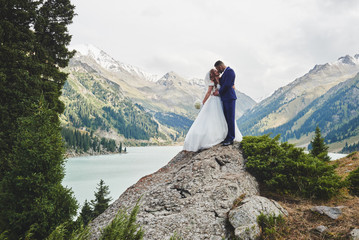 Beautiful wedding photo on mountain lake. Happy Asian couple in love, bride in white dress and groom in suit are photographed against background of the Kazakh landscape