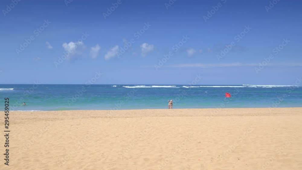 A tropical beach in hot afternoon with clear sand and couple is walking near blue water behind red flag swaying on wind A tropical beach in hot afternoon with clear sand and couple is walking near blue water behind red flag swaying on wind