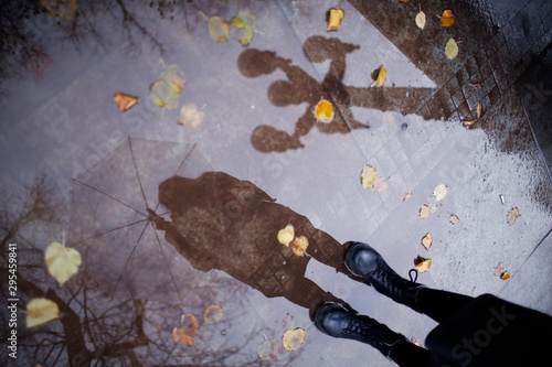 Girl in a coat, black shoes and with an umbrella. Autumn weather in the city, rain, reflection on the pavement. Drops of rain, fallen leaves.