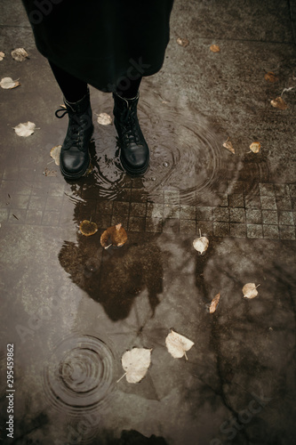 Girl in a coat, black shoes and with an umbrella. Autumn weather in the city, rain, reflection on the pavement. Drops of rain, fallen leaves.