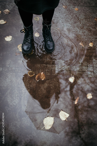 Girl in a coat, black shoes and with an umbrella. Autumn weather in the city, rain, reflection on the pavement. Drops of rain, fallen leaves.