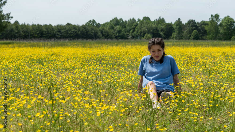 Teen girl sitting in large field with yellow flowers and trees in the background Teen girl sitting in large field with yellow flowers and trees in the background