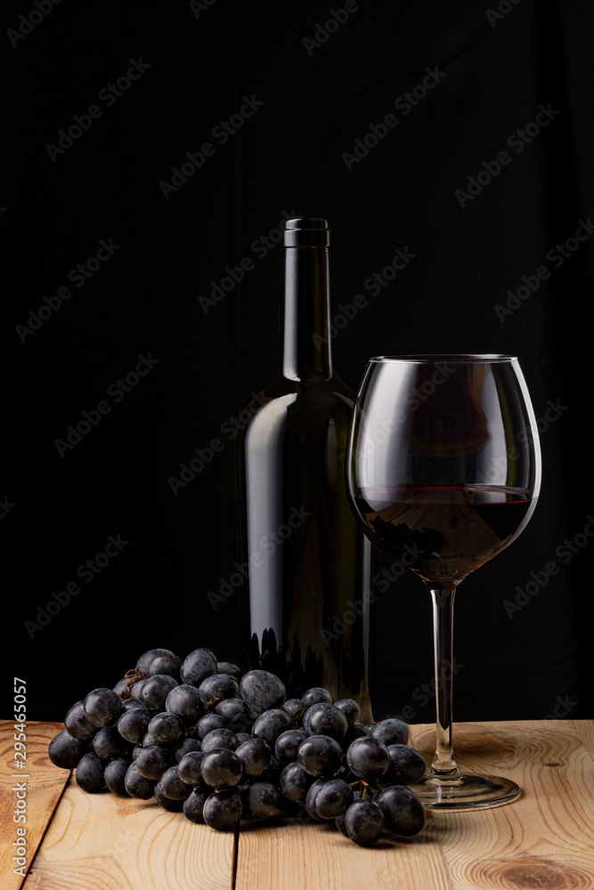Wine bottle and a glass of wine. A bunch of ripe black grapes. Wooden table. Black background.