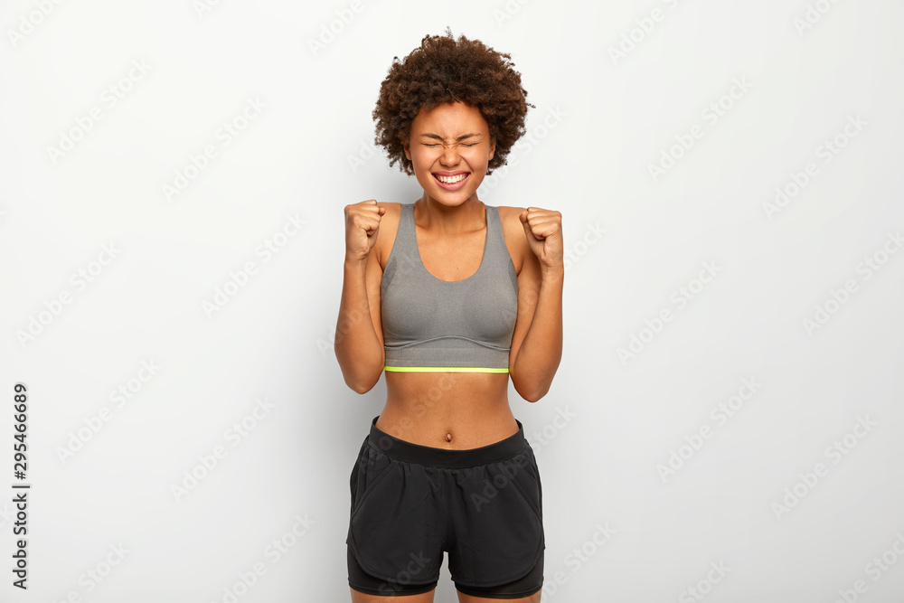 Energized sporty woman rejoices winning, raises clenched fists, smiles  broadly, wears sport bra , smiles broadly, isolated over white background,  awaits for something. People, triumph, success concept Stock Photo