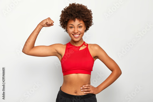 Tablou pe pânză Horizontal shot of positive dark skinned woman shows biceps, demonstrates strong hand, has slim figure, wears sport bra, smiles pleasantly, isolated over white background