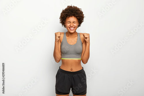Energized sporty woman rejoices winning, raises clenched fists, smiles broadly, wears sport bra , smiles broadly, isolated over white background, awaits for something. People, triumph, success concept