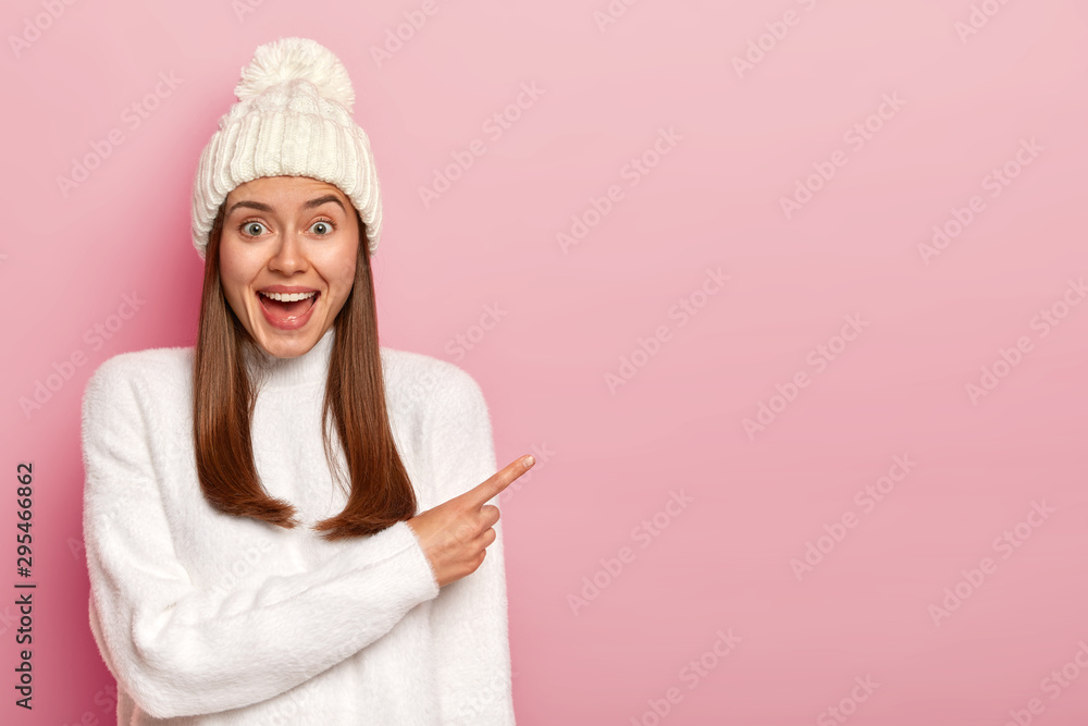 Amused cheerful woman points away with index finger, wears white hat and sweater, enjoys interesting scene, has long straight hair, suggests buying something, grins and shows copy space for promo - obrazy, fototapety, plakaty 