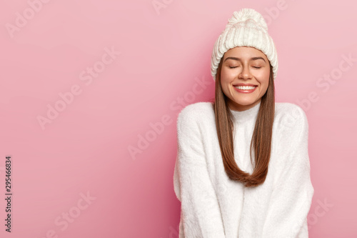 Horizontal shot of pretty young female with dark hair, keeps eyes shut, imagines pleasant moment, smiles pleasantly, shows white perfect teeth, enjoys comfort in new bought sweater, warm hat photo