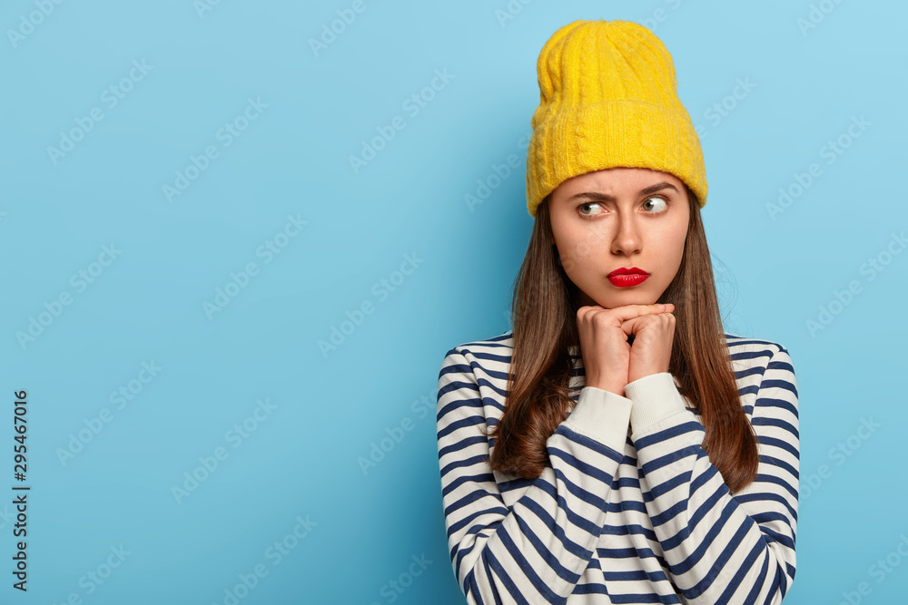 Studio shot of thoughtful young female model looks aside with serious expression, makes assumption, dressed in striped sweater, yellow stylish hat, isolated over blue background, blank space for promo Studio shot of thoughtful young female model looks aside with serious expression, makes assumption, dressed in striped sweater, yellow stylish hat, isolated over blue background, blank space for promo