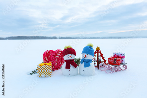 Couple two little snowman with the sledge, gift boxs, striped red white heart candy. Lawn covers with snow. Beautiful winter day. Happy new year 2020.
