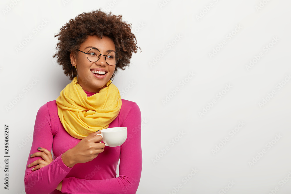Smiling glad dark skinned woman holds mug with aromatic coffee, has pleasant talk during drinking tea, wears optical glasses, yellow scarf and pink turtleneck, isolated over white background. Smiling glad dark skinned woman holds mug with aromatic coffee, has pleasant talk during drinking tea, wears optical glasses, yellow scarf and pink turtleneck, isolated over white background.