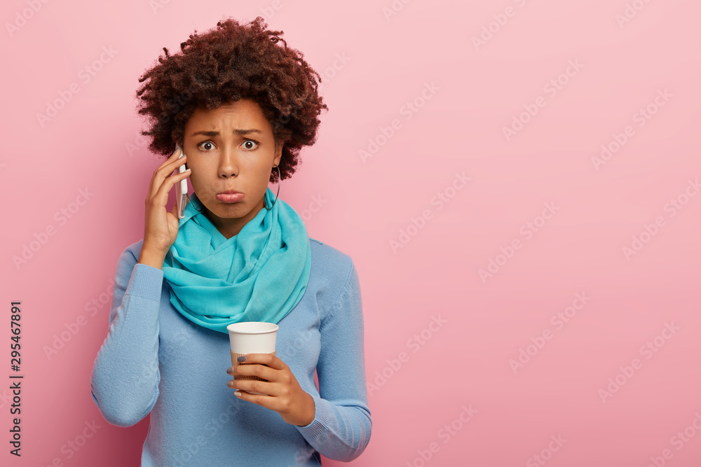 Studio shot of dissatisfied curly woman holds mobile phone near ear, has surprised unhappy look, drinks coffee, wears blue casual clothes, gets unpleasant news, poses against pink background Studio shot of dissatisfied curly woman holds mobile phone near ear, has surprised unhappy look, drinks coffee, wears blue casual clothes, gets unpleasant news, poses against pink background