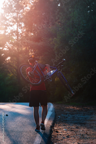 Strong athlete cyclist ( biker ) carrying his bicycle on his back on an asphalt road through deep green woods at sunset. Flat tire.