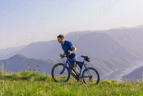 Tired fit mountain biker pushing his bike uphill at the top of the mountain on a sunny day with amazing view on a blue river.