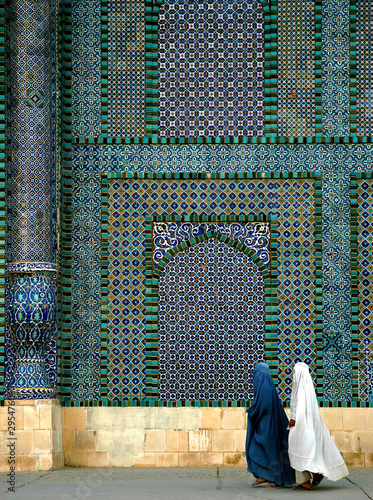 The Blue Mosque in Mazar-i-Sharif, Balkh Province in Afghanistan. Two women wearing burqas (burkas) walk past a wall of the mosque adorned with colorful tiles and mosaics. Northern Afghanistan. photo