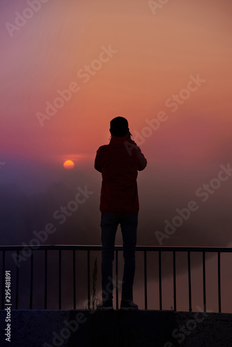 A silhouette of a man on a bridge over the river that photographs a sunrise on a foggy morning.