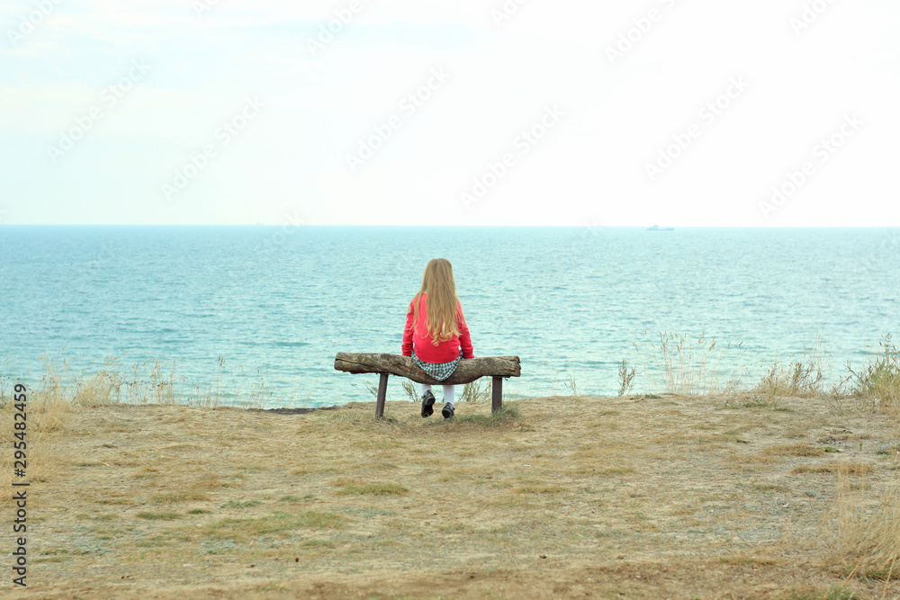 A little girl sits alone. Solo travelling. A tourist sits on a wooden bench on a cliff above the sea coast.