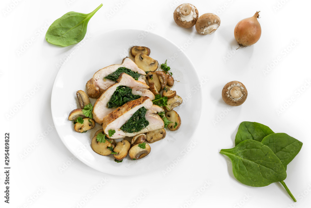 chicken breast fillet with spinach filling, mushrooms, onion and parsley garnish on a plate, some raw ingredients dispersed on the white background, copy space, high angle view from above chicken breast fillet with spinach filling, mushrooms, onion and parsley garnish on a plate, some raw ingredients dispersed on the white background, copy space, high angle view from above