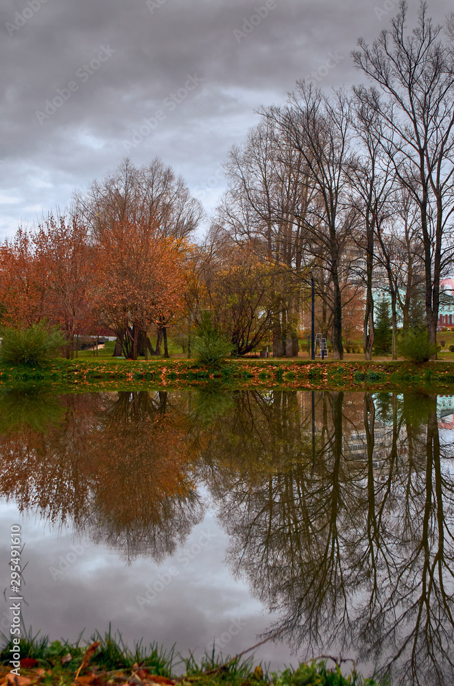 Trees with yellow and red leaves on the banks of the pond in the autumn park are reflected in the water. Trees with yellow and red leaves on the banks of the pond in the autumn park are reflected in the water.