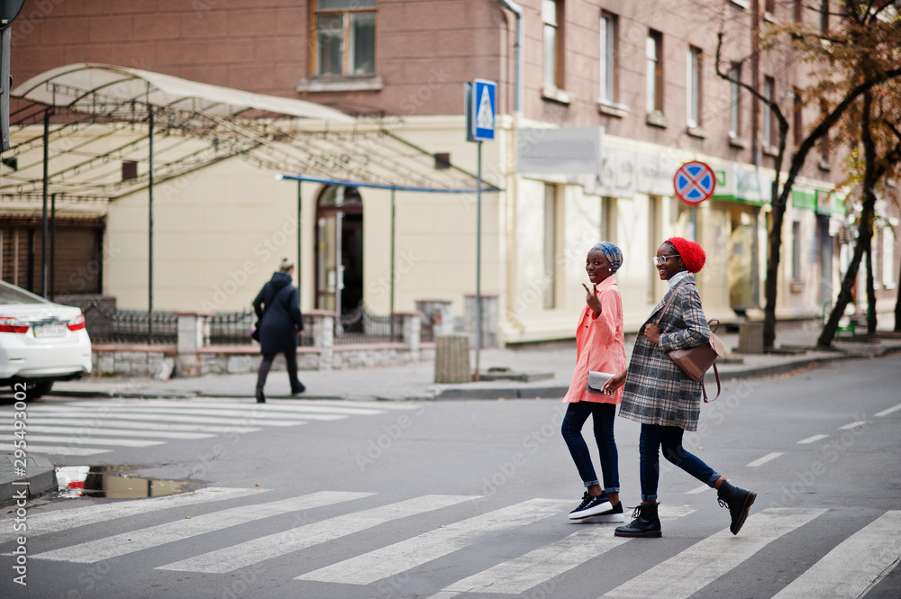 Two young modern fashionable, attractive, tall and slim african muslim womans in hijab or turban head scarf and coat walking on cross pedestrians.