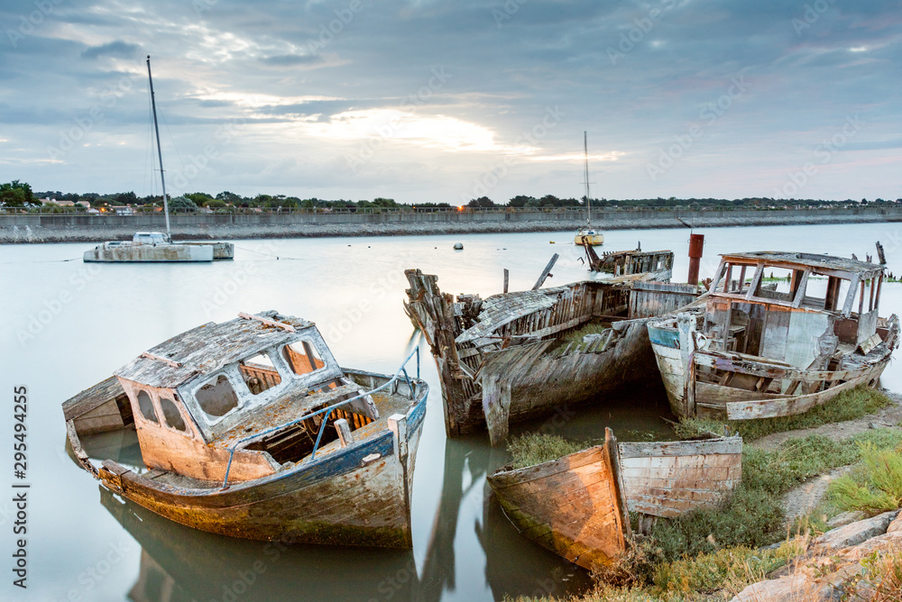The Noirmoutier boats cemetery early in the morning. A group of wrecks of old wooden fishing boats stranded in the mud is illuminated by the port lights. The Noirmoutier boats cemetery early in the morning. A group of wrecks of old wooden fishing boats stranded in the mud is illuminated by the port lights.