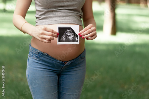 Happy young adult pregnant girl holds in her hands a snapshot of an ultrasonic study of a toddler walking in a park. Second trimester.
