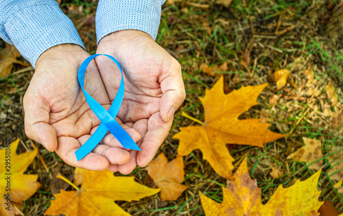 Men's health and prostate cancer awareness in November. Man hands holding a light blue ribbon awareness on the background of autumn  leaves. A symbol to support men who live with cancer.  Copy space.