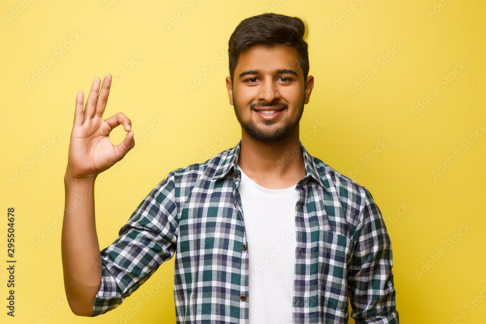 Young indian male looking like a tourist, okay sign concxept, isolated on yellow background. Young indian male looking like a tourist, okay sign concxept, isolated on yellow background.