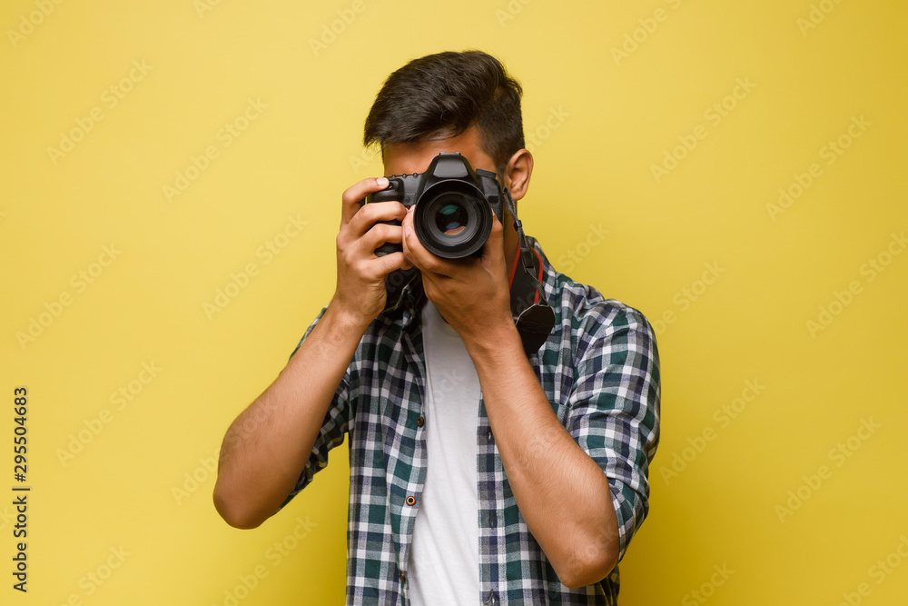 Handsome and confident indian man photographer with a large professional camera taking pictures photo shooting on the on the yellow background. - obrazy, fototapety, plakaty 