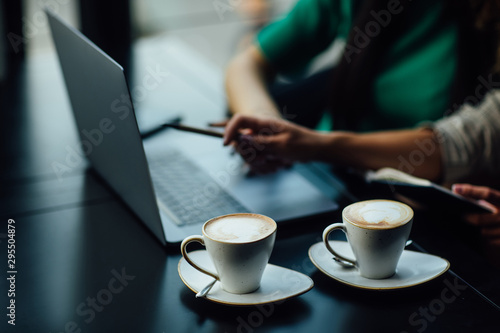 Close up photo. Girls sitting in a restaurant at the table, chatting and using a laptop while have a break time. Business concept, latte on table.