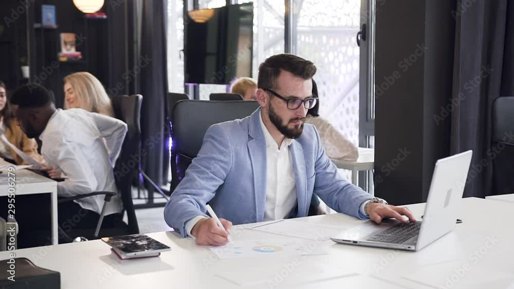 Good-looking experienced bearded office manager working with business documents and entering datas to laptop in the office room Good-looking experienced bearded office manager working with business documents and entering datas to laptop in the office room