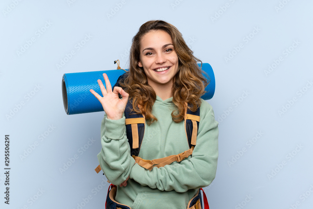 Young blonde hiker girl over isolated blue background showing ok sign with fingers - obrazy, fototapety, plakaty 