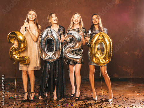 Beautiful Women Celebrating New Year.Happy Gorgeous Girls In Stylish Sexy Party Dresses Holding Gold and Silver 2020 Balloons  Having Fun At New Year s Eve Party.  arrying and drinking champagne flutes