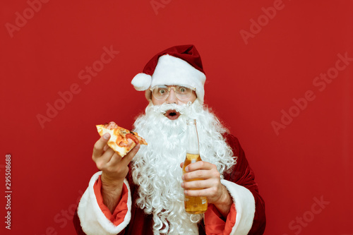 Surprised Santa Claus with piece of pizza and bottle of beer in hand stands on red background, looks into camera with shocked face, isolated. Funny Santa eats junk food and drinks alcohol.