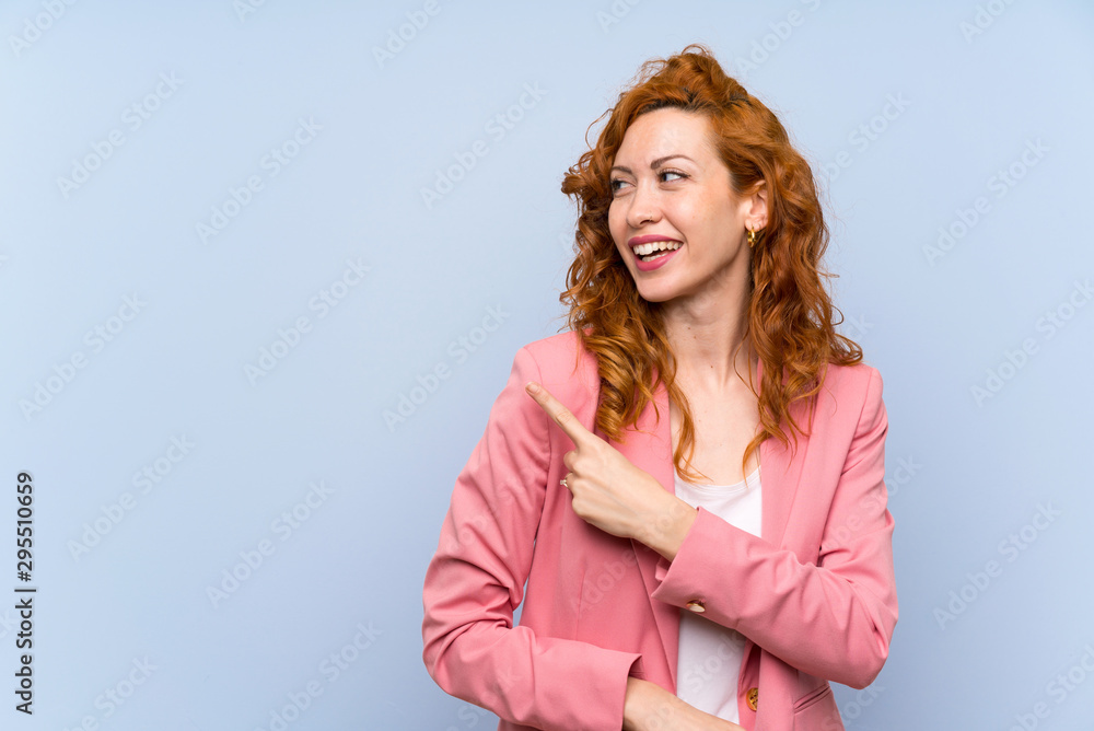 Redhead woman in suit over isolated blue wall pointing to the side to present a product Redhead woman in suit over isolated blue wall pointing to the side to present a product