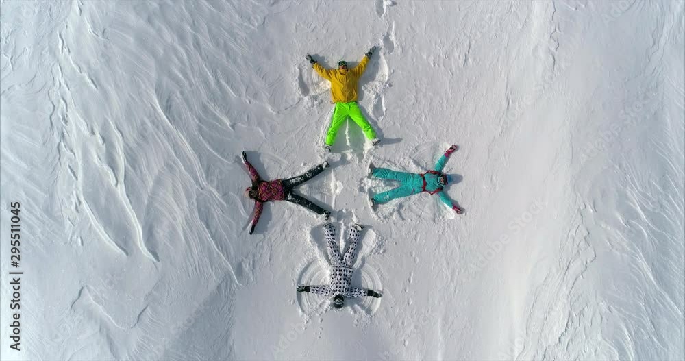 AERIAL TOP UP: a group of young people in bright ski suits depict snow angels lying on the fresh snow in the form of a cross. four winter angels happy and energetic with splashes of snow. SLOW MOTION