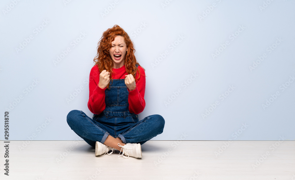 Redhead woman with overalls sitting on the floor frustrated by a bad situation Redhead woman with overalls sitting on the floor frustrated by a bad situation