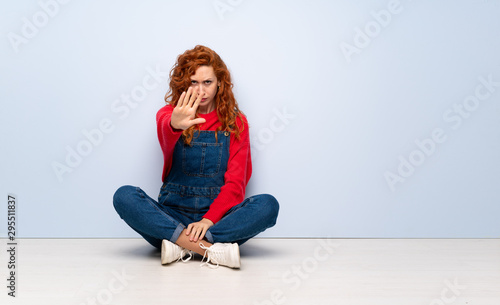 Redhead woman with overalls sitting on the floor nervous stretching hands to the front