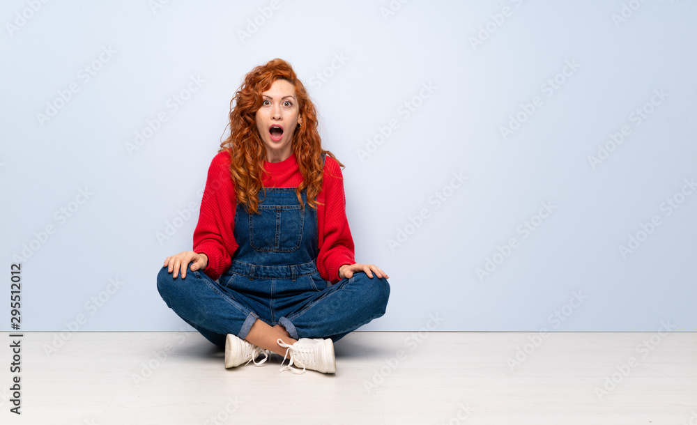 Redhead woman with overalls sitting on the floor with surprise facial expression Redhead woman with overalls sitting on the floor with surprise facial expression