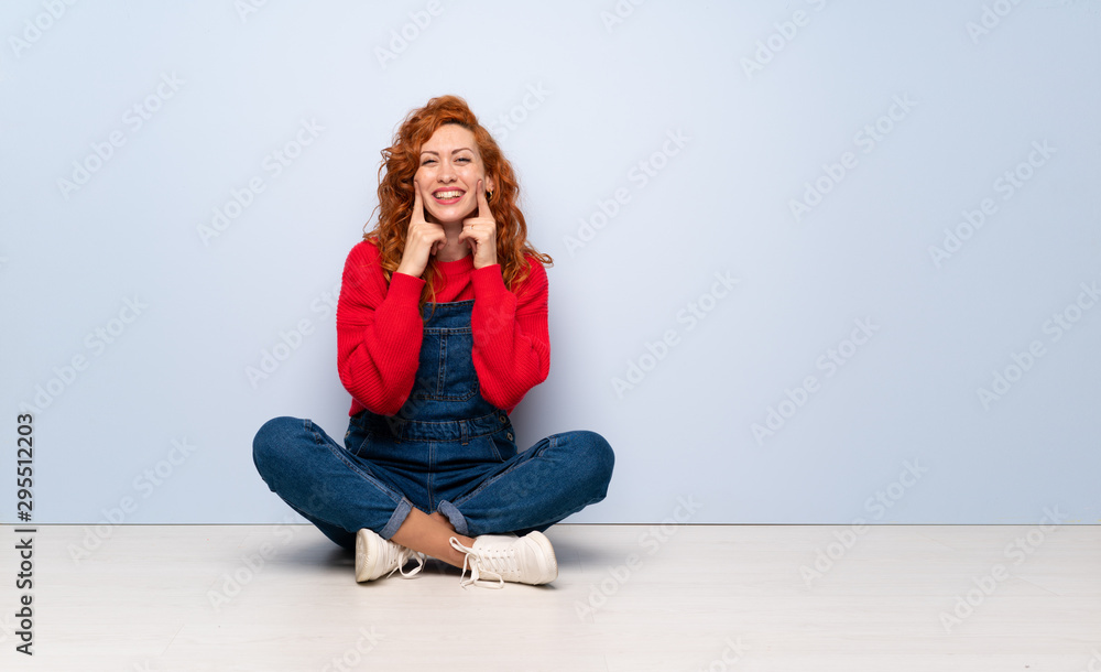Redhead woman with overalls sitting on the floor smiling with a happy and pleasant expression Redhead woman with overalls sitting on the floor smiling with a happy and pleasant expression