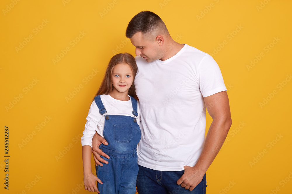 Close up portrait of pretty little girl andher dad, young father hugging her beautiful charming female kid, family looking at camera and smile while standing against yellow studio wall. Family concept Close up portrait of pretty little girl andher dad, young father hugging her beautiful charming female kid, family looking at camera and smile while standing against yellow studio wall. Family concept