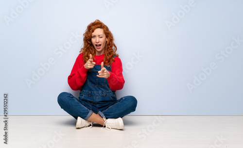 Redhead woman with overalls sitting on the floor pointing to the front and smiling