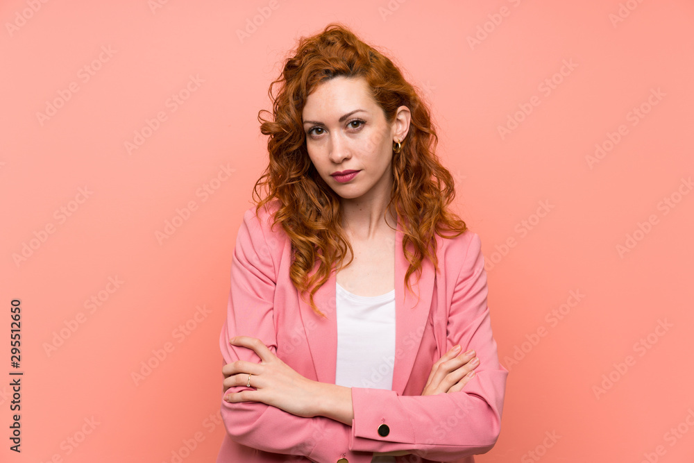 Redhead woman in suit over isolated pink wall keeping arms crossed Redhead woman in suit over isolated pink wall keeping arms crossed