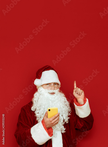 Santa Claus stands on red background with smartphone in hand, looks into camera with serious face and shows thumb up on copyspace. Isolated portrait. Vertical photo.