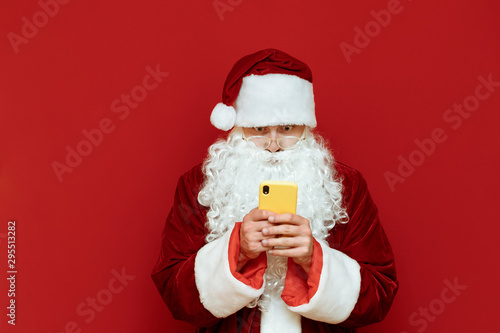 Portrait of shocked Santa Claus stands on red background with smartphone in hand and looks surprised at phone screen. Surprised Santa is using smartphone, isolated. X-mas