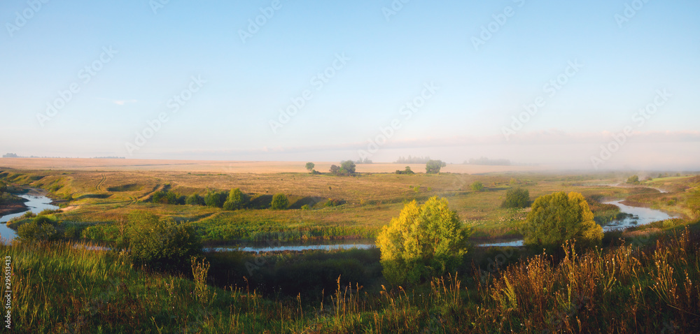 Hazy summer morning landscape with small river and golden fields