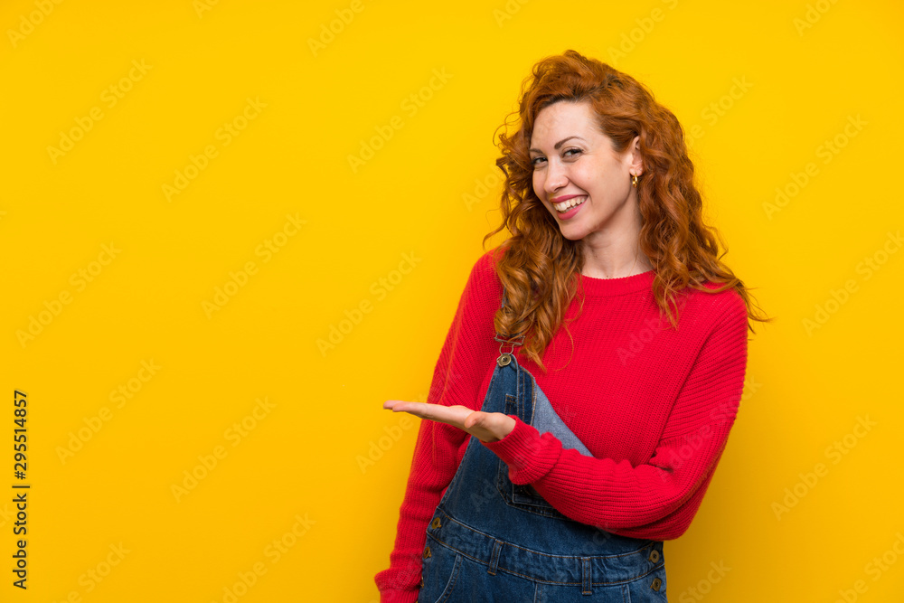 Redhead woman with overalls over isolated yellow wall extending hands to the side for inviting to come Redhead woman with overalls over isolated yellow wall extending hands to the side for inviting to come