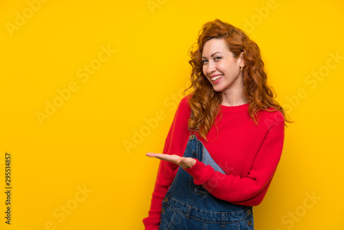 Redhead woman with overalls over isolated yellow wall extending hands to the side for inviting to come
